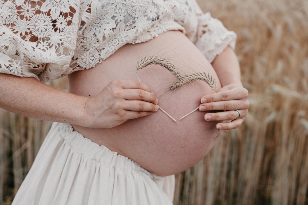 babybauch1 Schwangere Frau im weißen Kleid hält ein Herz aus Ähren vor ihrem Babybauch in einem Weizenfeld.