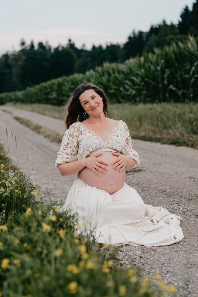 babybauch10 Schwangere Frau sitzt im weißen Kleid auf einem Feldweg und hält ihren Babybauch mit beiden Händen.