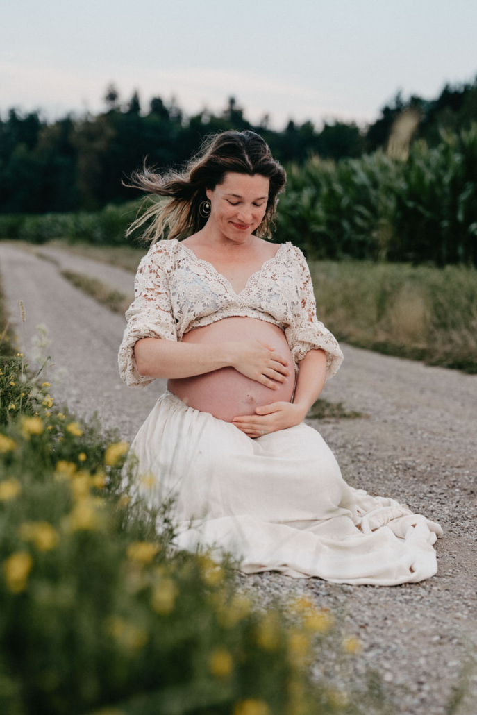 babybauch15 Schwangere Frau sitzt im weißen Kleid auf einem Feldweg und hält ihren Babybauch mit beiden Händen.