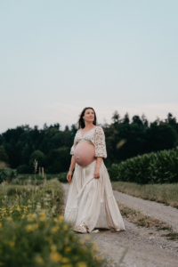 Schwangere Frau steht auf einem Feldweg und hält ihr fließendes Kleid während eines Babybauchshootings in der Natur.