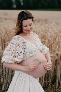 Schwangere Frau im weißen Kleid hält ein Herz aus Ähren vor ihrem Babybauch in einem Weizenfeld.