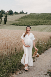 Schwangere Frau steht auf einem Feldweg und hält ihr fließendes Kleid während eines Babybauchshootings in der Natur.