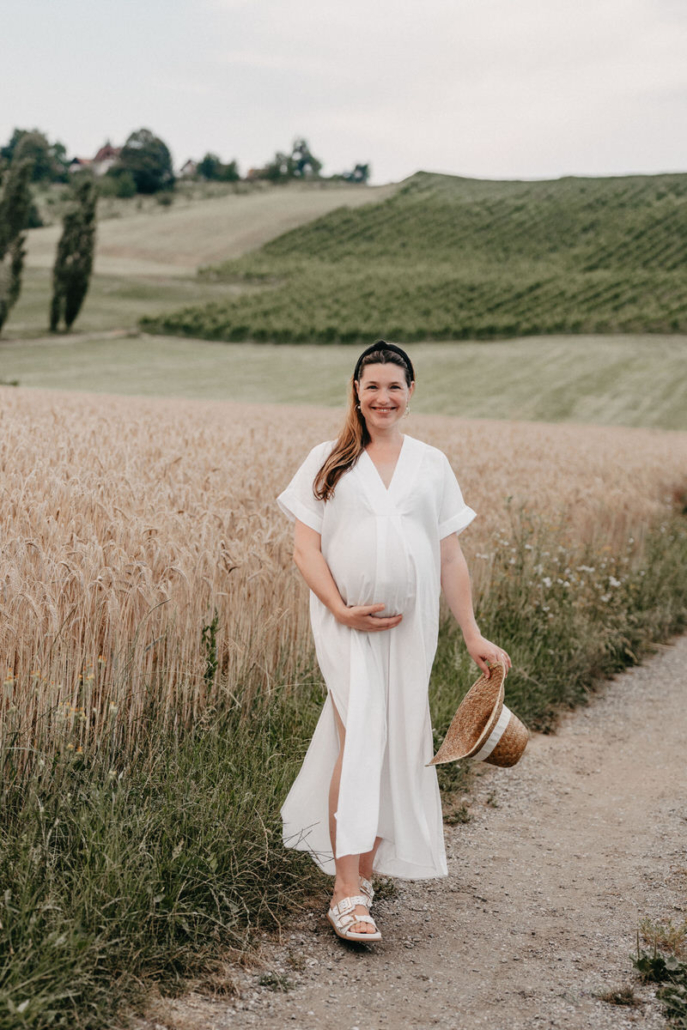 babybauch30 Schwangere Frau steht auf einem Feldweg und hält ihr fließendes Kleid während eines Babybauchshootings in der Natur.