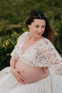 Schwangere Frau sitzt im weißen Kleid auf einem Feldweg und hält ihren Babybauch mit beiden Händen.