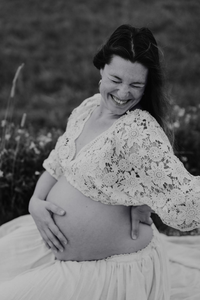 babybauch8 Schwangere Frau sitzt im weißen Kleid auf einem Feldweg und hält ihren Babybauch mit beiden Händen.